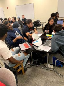 Small groups of students seated around tables, talking and working on their laptops.